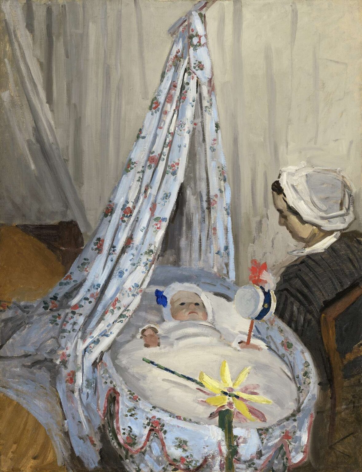 A woman dressed in dark clothing and white cap sits beside a covered cradle draped with floral fabric; an infant lies inside, gazing upward at suspended toys under soft, diffused light.