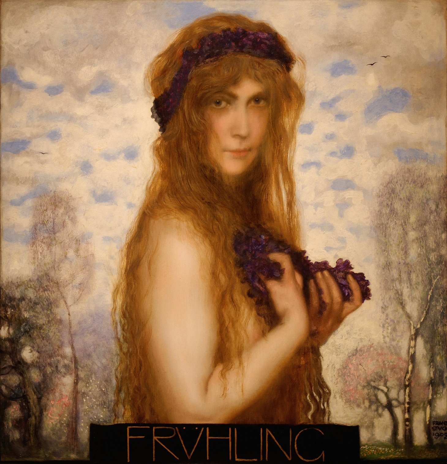 A serene woman with flowing auburn hair, crowned with purple flowers and holding a floral garland, gazes softly at the viewer against a dreamy springtime sky in Franz von Stuck’s 1902 painting The Spring.