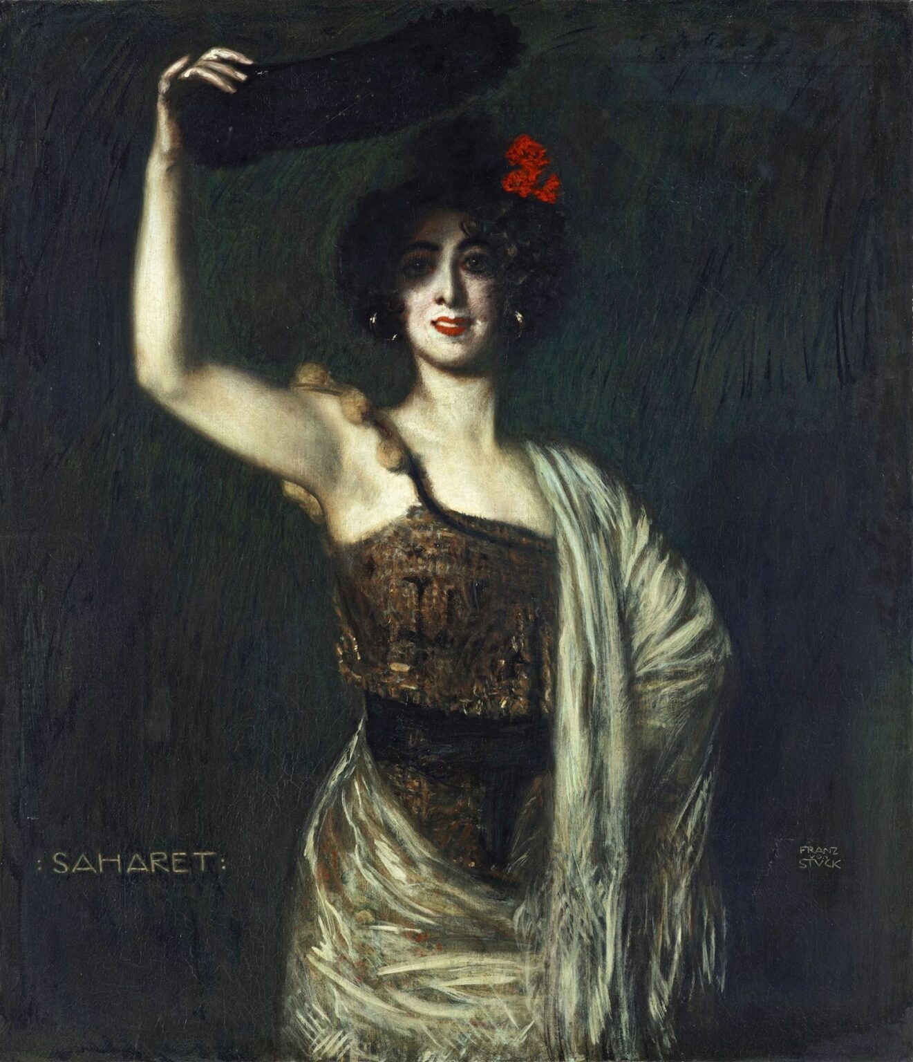 Portrait of dancer Saharet standing in a dark green‑black space, raising a fan above her head, wearing a velvet bodice and ivory fringed shawl, with a red flower in her curly hair.