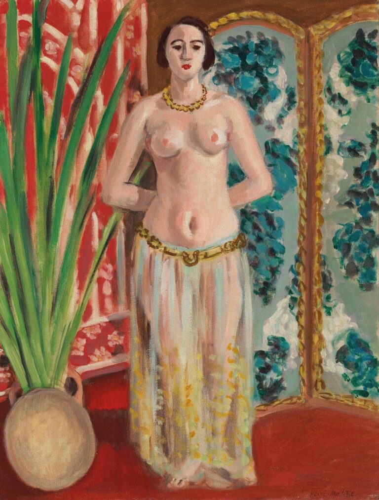 A half-nude woman stands frontally on a red floor with her hands behind her back, wearing a gold necklace and belt with a translucent white skirt. At left a pot holds tall green leaves; at right a folding screen with blue blossoms and gold trim forms the backdrop, with a red patterned fabric on the left wall. Painted by Henri Matisse in 1923.