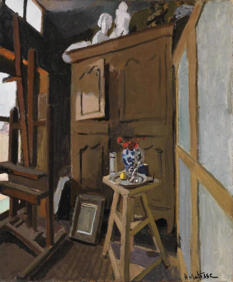 Sunlit artist’s studio with a dark window and easel at left, large olive-brown armoire at center topped by white plaster studies, a stool-table holding a patterned vase, tin, cup, dish, and lemon, pale panelled wall at right, and a leaning empty frame on the floor; painted with clear planes and restrained color.