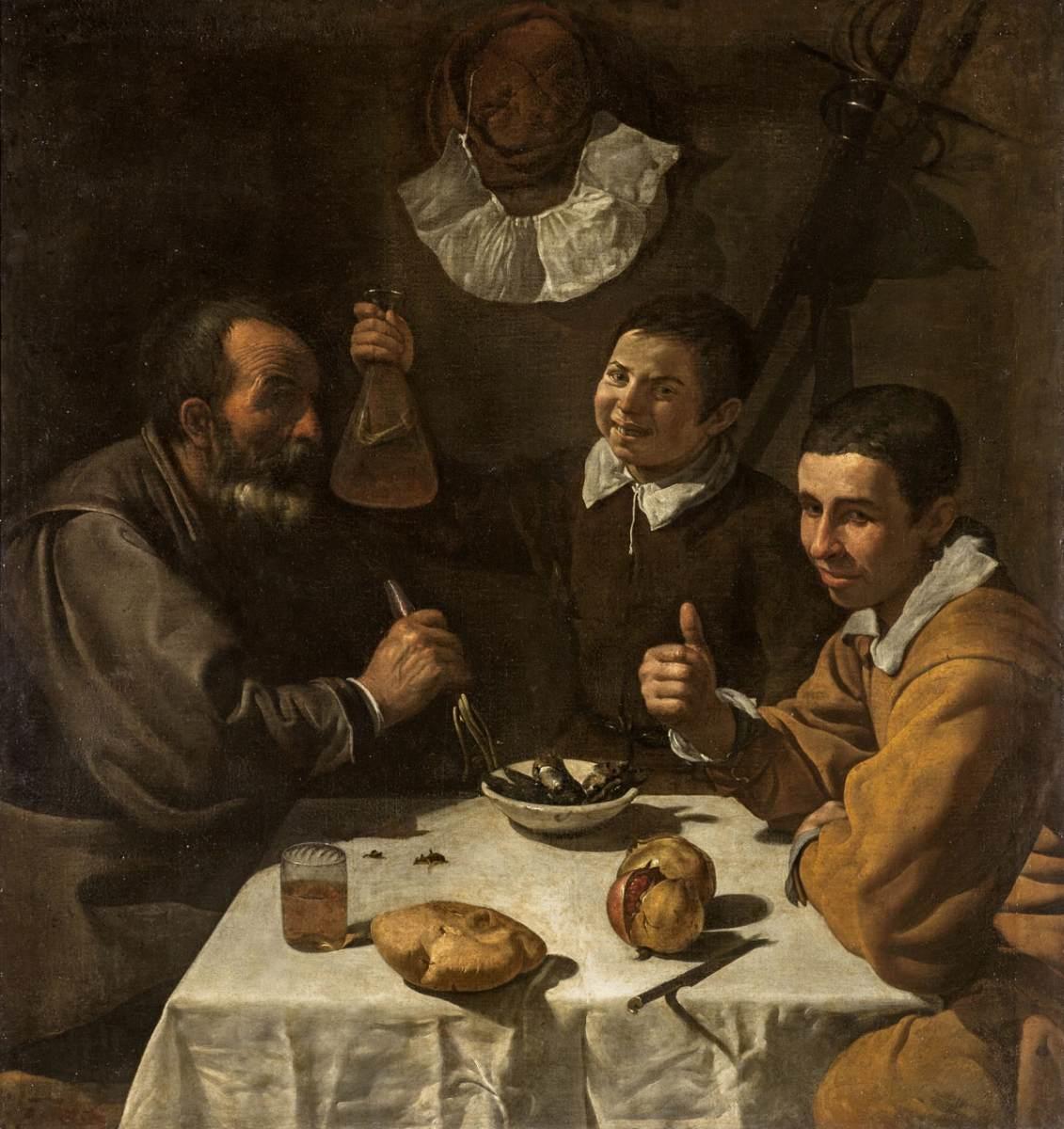 Three figures sit at a small table under strong light: an older man with a fork, a boy raising a glass carafe, and a youth giving a thumbs-up. On the white cloth lie a loaf, a split pomegranate, a glass of wine, a bowl of small fish, and a knife, all set against a dark background.