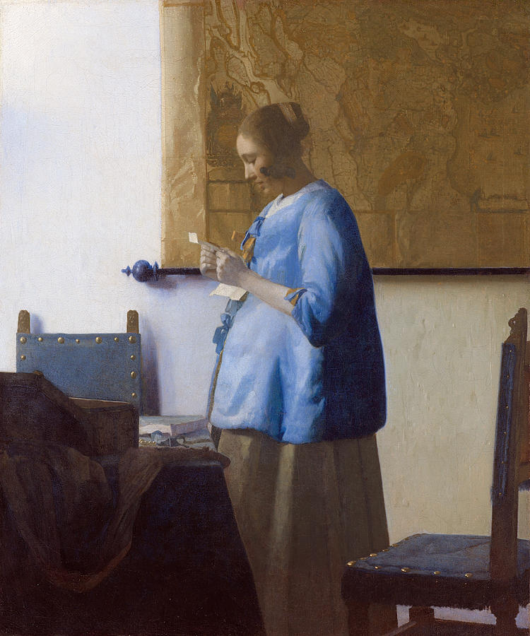 A young woman in a deep blue jacket stands in profile reading a letter beside a table and two studded chairs, while a large ochre wall map hangs behind her in a softly lit, sparsely furnished Delft room.
