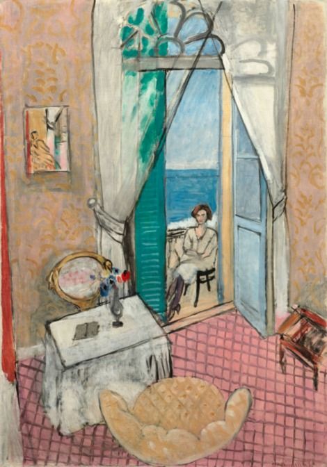 Tall, sunlit room with pink tiled floor and patterned ochre walls; a table with brushes, scalloped armchair, and sheer curtains frame a central balcony door where a woman sits facing a blue sea under pale sky; mint shutters and delicate tracery complete the opening.