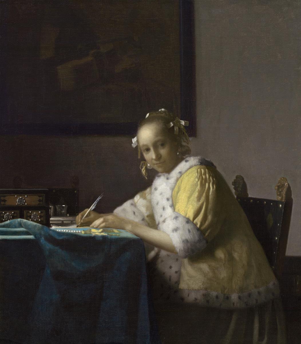 A woman in a yellow fur-trimmed jacket sits at a blue-covered table holding a quill over a sheet of paper, with pearls, a small casket, and a glass nearby, in a softly lit Delft room with a dark painting on the wall behind her.
