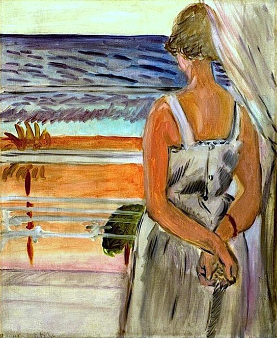 A woman seen from behind stands beside a pale curtain and looks out to a Mediterranean view of ochre beach, turquoise shallows, and deep blue sea; her grey dress and clasped hands anchor the interior in Matisse’s Nice-period style.