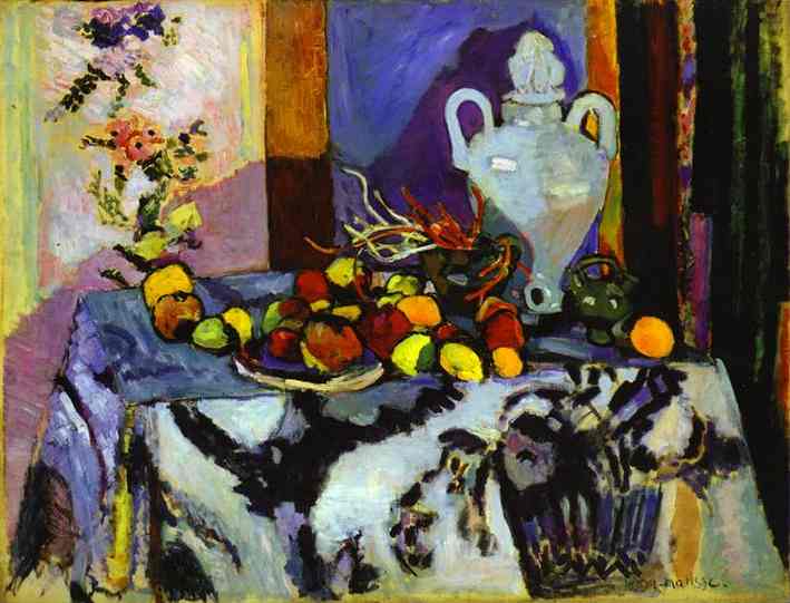 A richly colored still life by Henri Matisse showing a table draped with a bold patterned blue cloth, piled with fruit, flanked by small green vessels, and dominated by a pale urn against lavender and multicolored wall panels, painted with confident brushwork and luminous color.