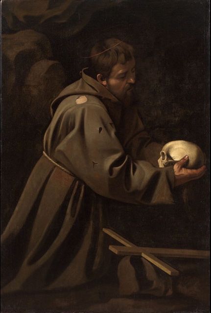 In a dark cave-like setting Saint Francis kneels in a patched brown habit, gently holding a skull in both hands while a rough wooden cross lies on the ground; a narrow beam of light illuminates his face, sleeves, and the ivory skull against deep shadow.