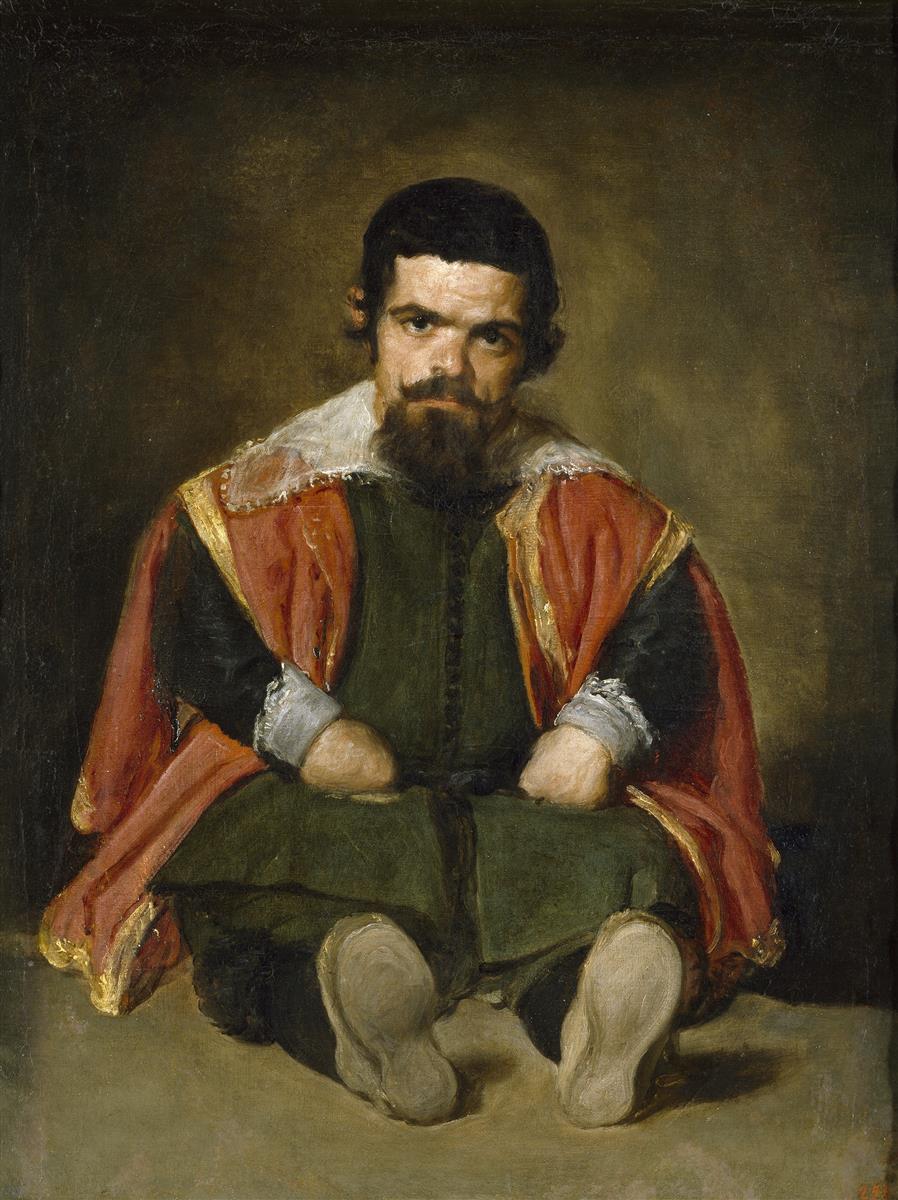 A man with dwarfism sits frontally on the floor in a red, gold-trimmed mantle over a dark green outfit, hands beside his knees and feet forward, looking directly at the viewer against a softly dark background.