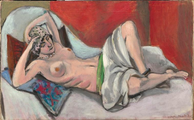 Reclining semi-nude woman on a pale couch, head in a dotted scarf, necklace at the throat; a cool gray-white drape crosses her hips with a green waistband accent; patterned blue pillow at left; large red screens form the background, painted in soft, luminous tones typical of Matisse’s Nice period.