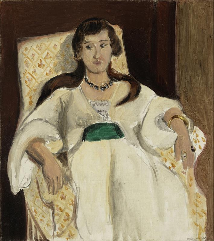 A woman in a flowing white dress sits in a patterned armchair, a deep green sash at her waist, with dark hair, a bead necklace, and relaxed hands; warm browns frame the shallow interior, painted with broad luminous strokes.