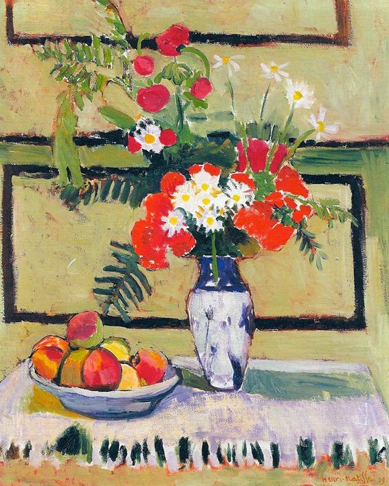 A pale yellow-green paneled wall frames a white-fringed table holding a blue-patterned vase of red and white flowers with green ferns beside a shallow bowl of peaches; bold dark contours and vivid planes organize the still life in Matisse’s 1909 “Flowers and Fruit.”