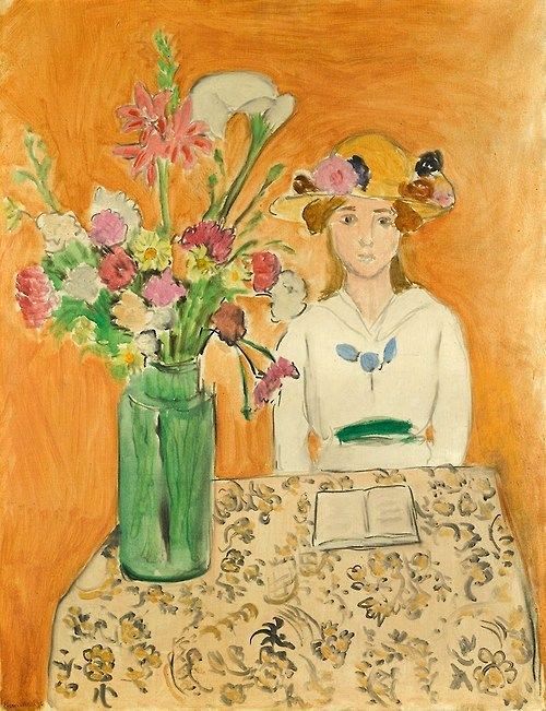 A girl in a white dress and flowered straw hat sits at a patterned table with an open book; to her left a tall green vase holds a colorful bouquet; the background is a warm orange wall, painted with loose, luminous brushstrokes.
