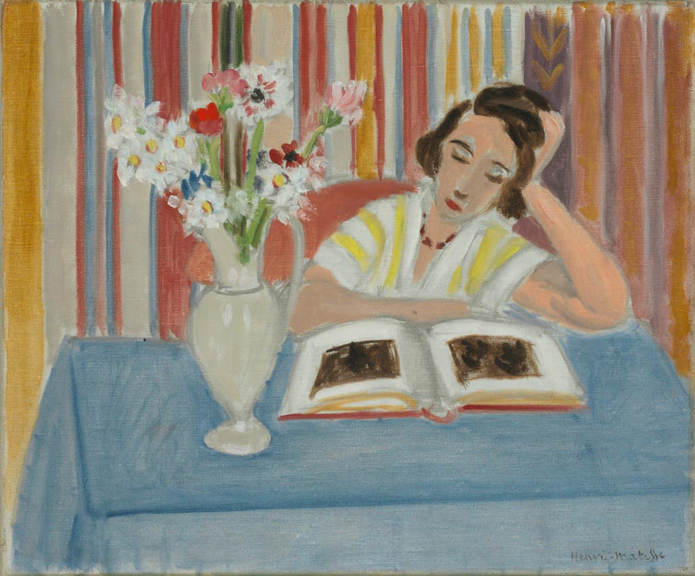 Calm interior with a woman reading at a table covered in blue cloth; a pale ceramic vase of mixed flowers stands to her left; behind her hangs a curtain of vertical multicolored stripes; soft light, gentle colors, and simplified forms characteristic of Matisse’s Nice period.