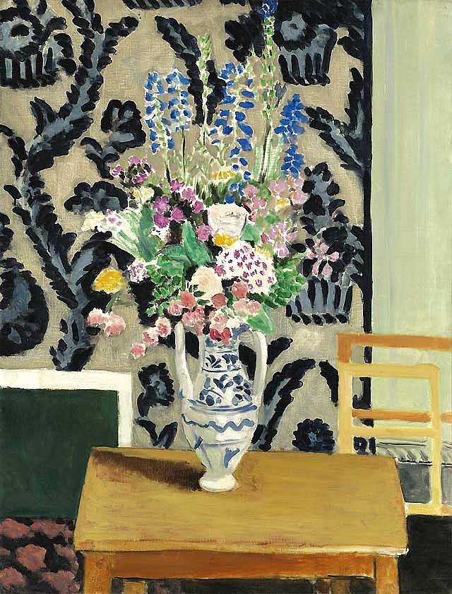 A tall bouquet of pink, white, and blue flowers in a blue-and-white vase sits on a wooden table before a beige wall covered with bold dark leaf patterns, with a simple chair at the right edge.