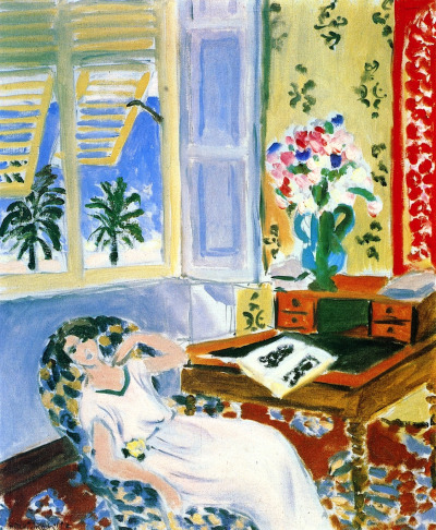 Sunlit room with open yellow shutters revealing blue sea and palms; a woman in a pale dress naps on a patterned armchair; at right a wooden desk holds an open book and a green vase of pink and blue flowers; floral carpet and wallpaper unify the airy Nice-period interior.