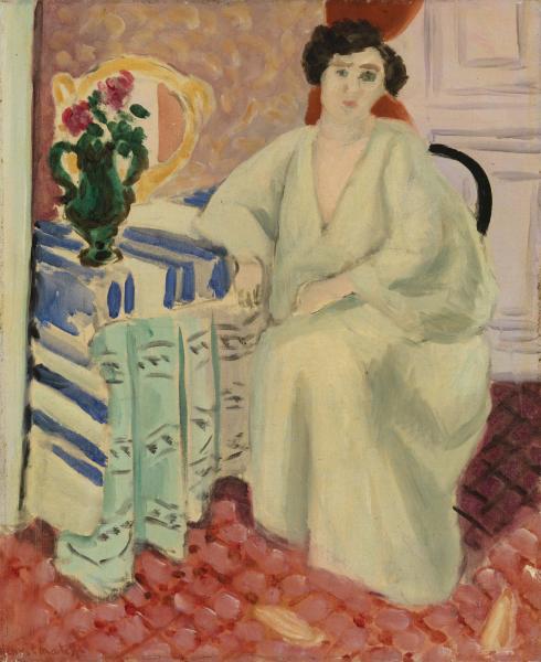 A woman in a pale robe sits beside a blue-and-white striped table holding a green jug of flowers and a round golden mirror; a coral patterned carpet covers the floor and a paneled door and chair back appear at right, in a Nice-period interior by Henri Matisse.