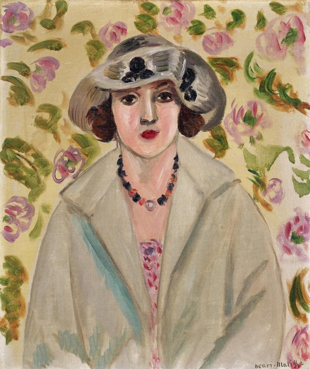 Frontal portrait of a young woman wearing a wide white horsehair hat and pale coat, a bead necklace at her neck, and a glimpse of patterned dress; behind her a cream wall scattered with pink roses and green leaves; soft, even light and restrained colors characteristic of Matisse’s Nice period.