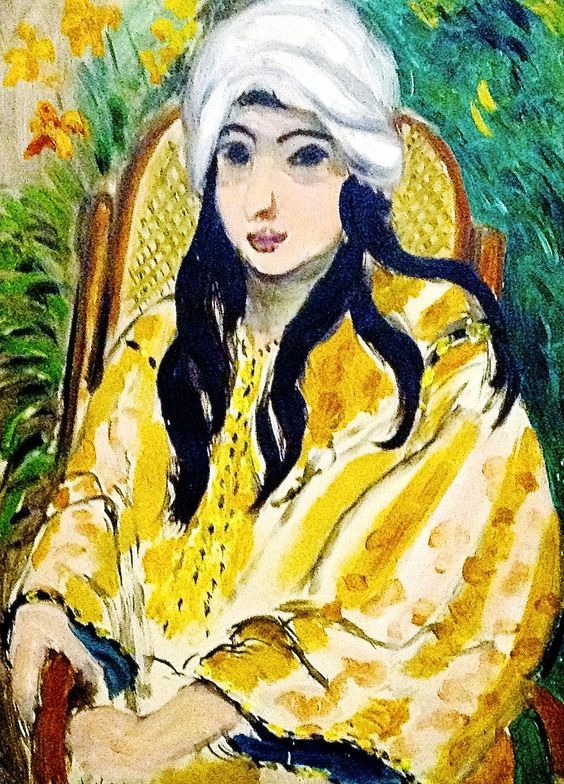 A seated young woman painted by Matisse wears a bright white turban and flowing yellow robe, her long black hair falling in curves over the garment; she sits in a cane-backed chair against a shallow background of green foliage, with strong black contours and broad, lively brushstrokes creating a calm, modern portrait.
