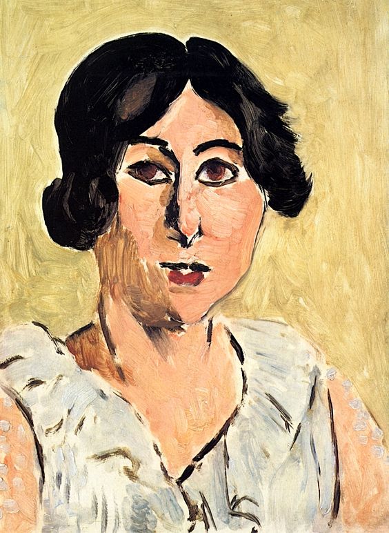 A close, bust-length portrait by Matisse of a woman with dark hair and strongly outlined black eyes, set against a warm ochre background; her pale dress is painted in cool whites and grays, and bold black contours define features and silhouette with simplified planes and visible brushstrokes.