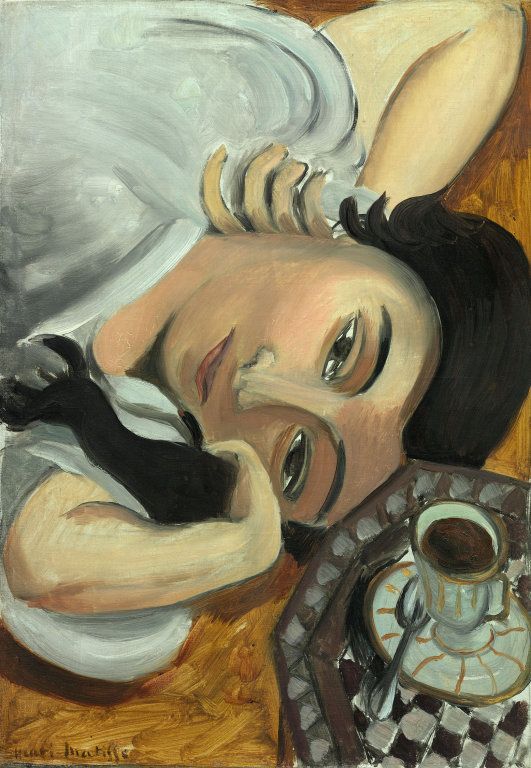 A close, diagonal portrait by Matisse of Lorette lying on her side with her cheek on her forearm; her dark hair contrasts with pale skin and a cool white blouse, while a small tray with a demitasse and saucer sits at the lower right on a checkered surface; warm wood tones, gentle light, and strong black contours create a quiet, intimate scene.