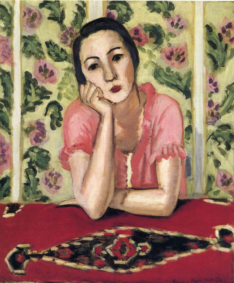 A woman in a coral-pink blouse rests her chin on her hand at a red table with a dark medallion pattern; behind her, vertical panels of pale wallpaper bloom with green leaves and rose flowers, all rendered in soft, even light that gives the portrait a serene, decorative atmosphere.