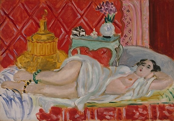Nude woman reclining on a divan draped in white before a quilted red wall; turquoise side table with bouquet and small objects; large gold vessel at left; shallow, decorative interior painted by Henri Matisse in 1926.
