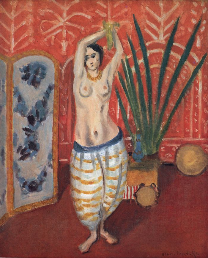 A topless woman in loose striped trousers stands with arms raised in a red, patterned interior; a tall green plant fans behind her, a blue-panel folding screen sits to the left, and tambourines rest on the floor as warm colors and bold shapes create a balanced, decorative scene.