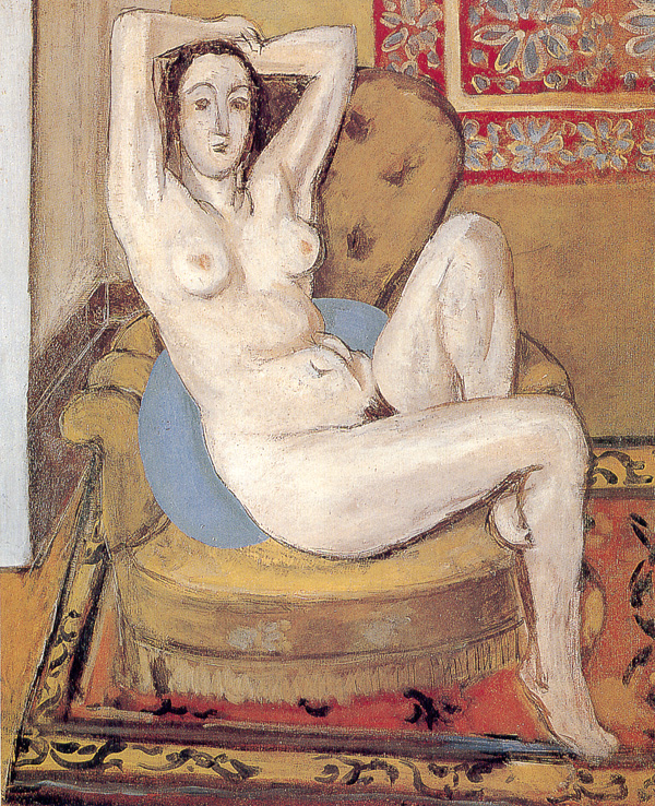 A pale nude reclines in a golden armchair with one arm raised and one knee bent; a round blue cushion supports her back; a patterned carpet and a floral wall textile frame the scene, creating a calm, decorative interior bathed in soft light.