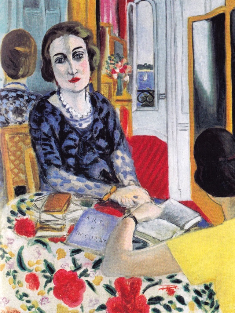 A woman with a pearl necklace sits at a floral-covered table facing the viewer; her image is also seen in a mirror behind her alongside a bouquet and a glimpse of blue sea. A companion in a yellow dress is seen from behind, hands meeting near open books on the table, all within a brightly colored, patterned interior.