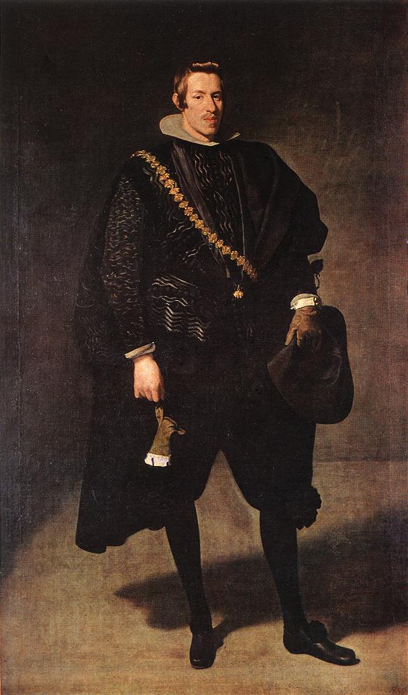 Full-length portrait of Infante Don Carlos standing in a quiet gray interior, dressed in black court attire with a high white collar and diagonal gold chain, holding a broad-brimmed hat in one hand and a glove in the other as light from the left models his face and clothing.
