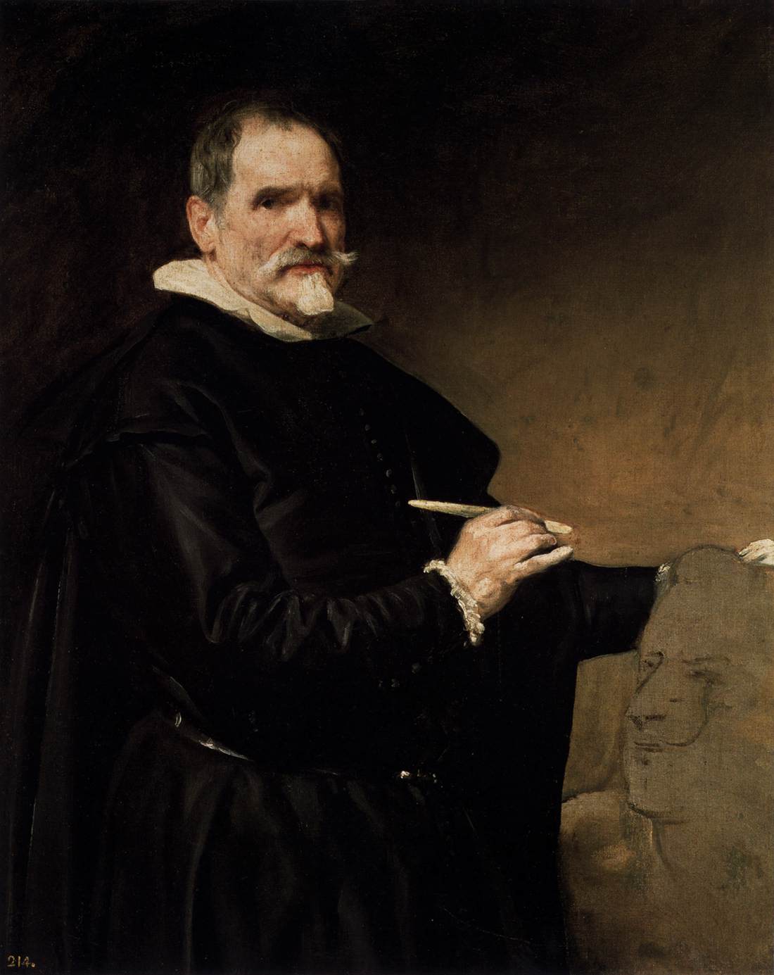 Three-quarter portrait of sculptor Juan Martínez Montañés in black attire with white collar, holding a modeling tool as he works on a clay head; his illuminated hand and thoughtful gaze stand out against a warm brown studio background.