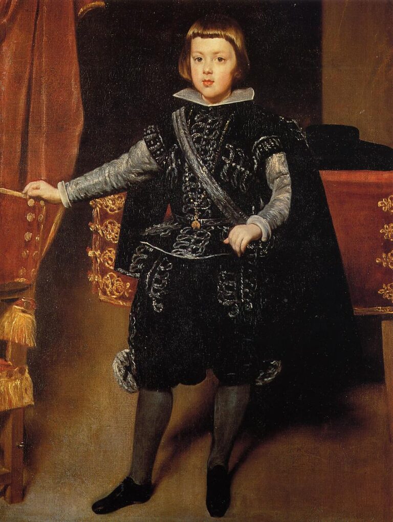 Full-length portrait of young Prince Balthasar Carlos standing between a gilded chair and red-covered table, wearing black velvet embroidered with silver, white lace collar and cuffs, and a short cape, facing forward under warm interior light.