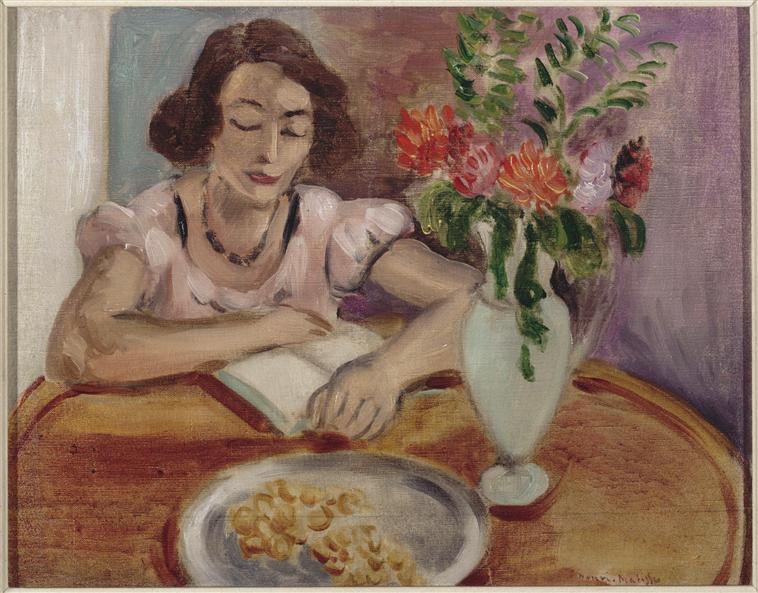 Interior with a woman in a pink dress reading at a round wooden table; a pale vase of orange-coral flowers and green leaves stands to her right; a silver plate with small pastries sits in the foreground; soft lavender wall and warm wood tones create a serene Nice-period atmosphere.