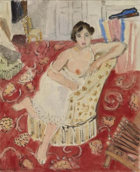 Interior with a woman in a white chemise reclining on a cream-patterned seat, surrounded by a richly patterned red carpet; pale gray back wall, small chair in distance, green cushion at left, and a bundle of wooden slats near the lower right.