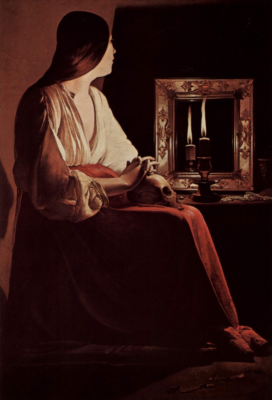 Candlelit interior with the Magdalene seated in profile, hands folded over a skull on her lap; on a table a single candle burns before a framed mirror that reflects a second flame, with pearls and a rosary resting in shadow.