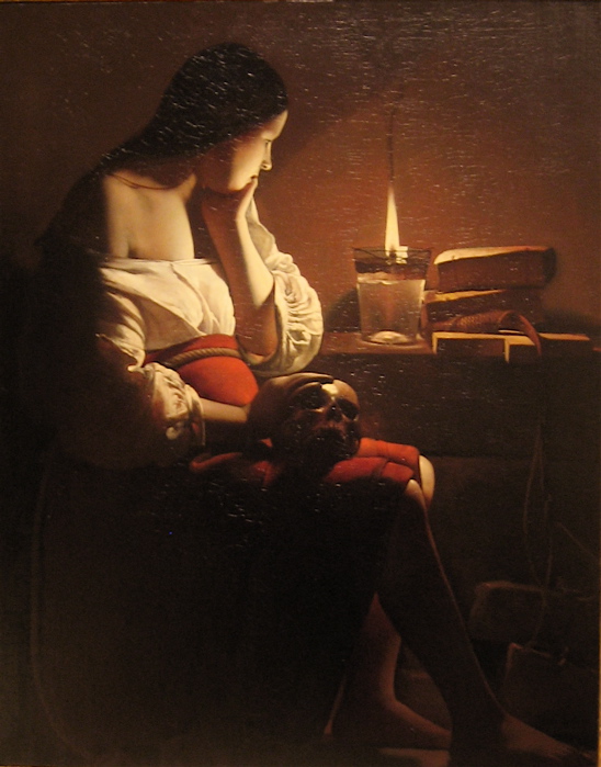 Mary Magdalene sits in profile in a dark room, her cheek resting on her hand beside a clear oil lamp; a skull rests on her lap, closed books and a coiled cord sit on a table, and warm lamplight models her white blouse and bare shoulder against a simple brown wall.