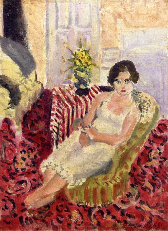 A woman in a pale dress reclines in a green striped armchair within a red patterned carpeted room; a small table with a red-and-white striped cloth and yellow flowers stands beside her, and luminous doors open in the background, painted by Henri Matisse in 1920.