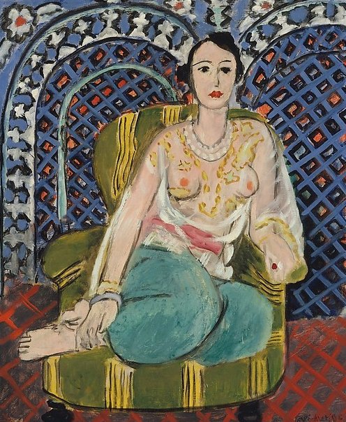 Seated woman with turquoise trousers and a sheer floral blouse sits cross-legged on a green striped armchair before a richly patterned blue screen; red tiled floor in foreground and simplified facial features typical of Matisse’s 1926 Nice interiors.