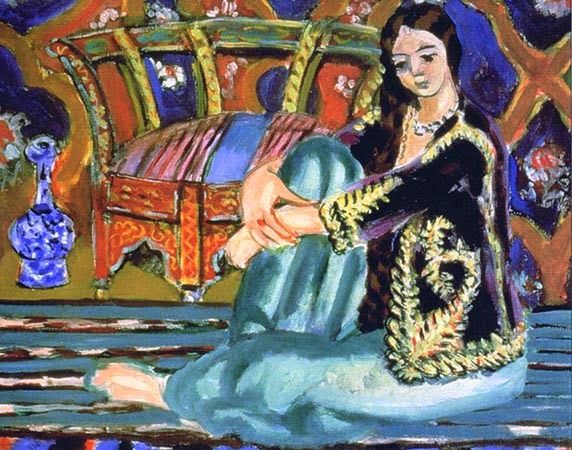 Seated woman with knees drawn up, wearing turquoise trousers and a black jacket with gold embroidery; pearls at her neck; behind her a curved red-orange bench and a violet-orange ornamental wall; blue patterned bottle at left and broad striped floor in the foreground; Henri Matisse, 1928.