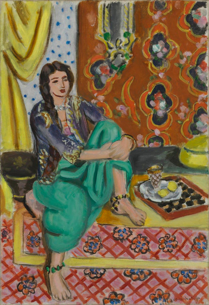 A woman in green trousers and a dark embroidered jacket sits cross-legged on a patterned carpet; behind her hang a lemon curtain, a dotted wall, and a russet tapestry with large motifs. At right a tray holds a small checkerboard, a tulip glass, and two lemons, all bathed in soft daylight.