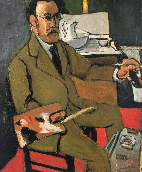 Matisse seated in an olive suit holding a palette and brush, red floor and chair, pale tabletop still life with white pitcher and bowl behind him, black accents outlining forms; shallow space and tuned color characteristic of his 1918 Nice-period style.
