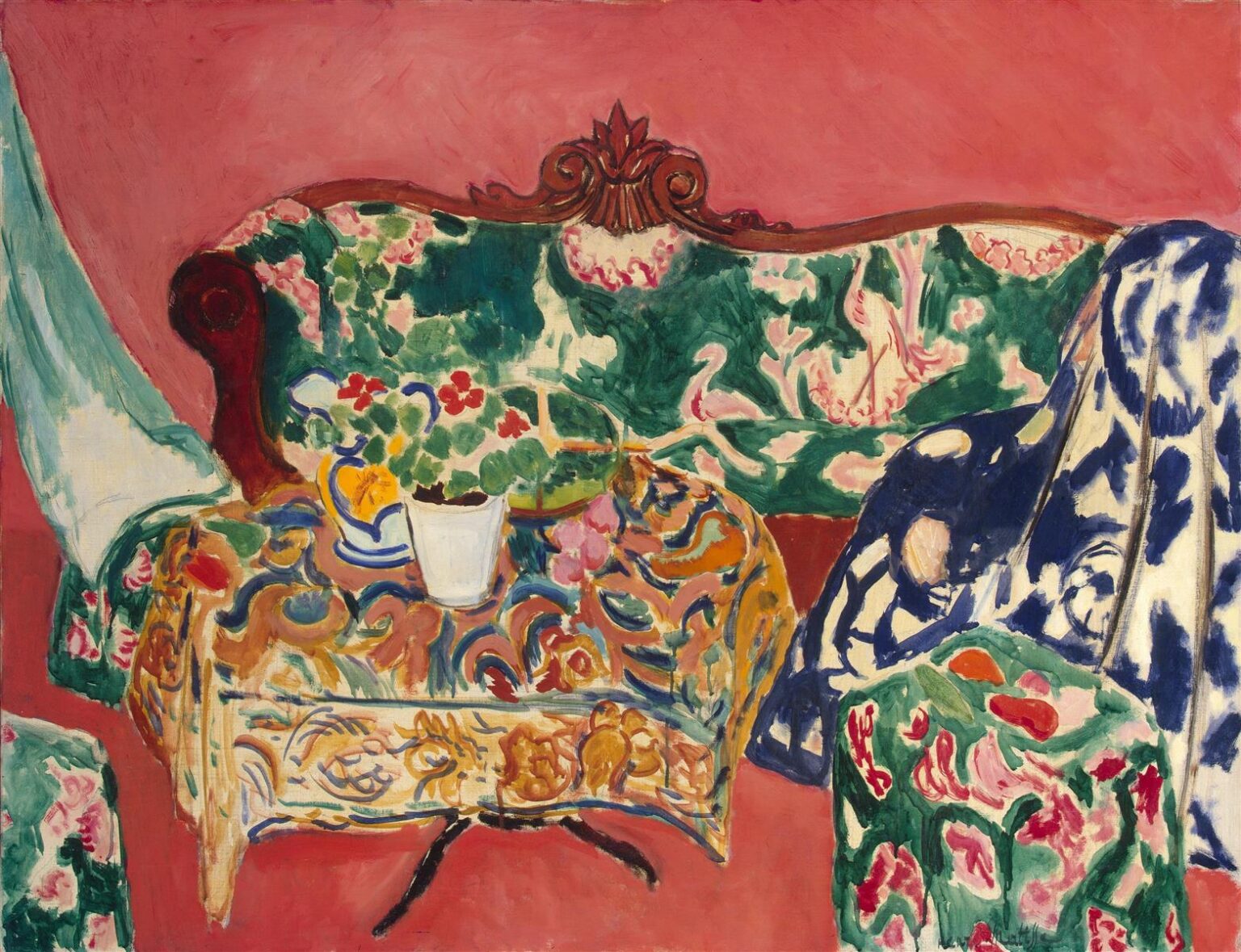 Coral-pink room with a carved green sofa upholstered in a swan-patterned fabric, an oval table draped in ocher and blue arabesque cloth holding a white pot of red and green flowers and a yellow-blue jug, a bold blue-and-white spotted shawl cascading at right, and a small floral ottoman in the foreground; all rendered with thick brushstrokes and strong complementary colors in Henri Matisse’s 1911 “Seville Still Life.”