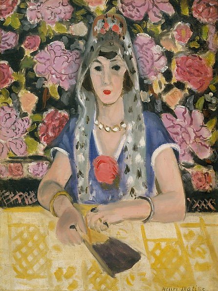 Frontal portrait of a woman seated at a table, wearing a blue dress, veiled headdress, and gold jewelry; a red rose at her chest; hands holding a small fan over a yellow patterned tablecloth; dense pink floral backdrop; overall harmony in blue with soft Nice-period light.