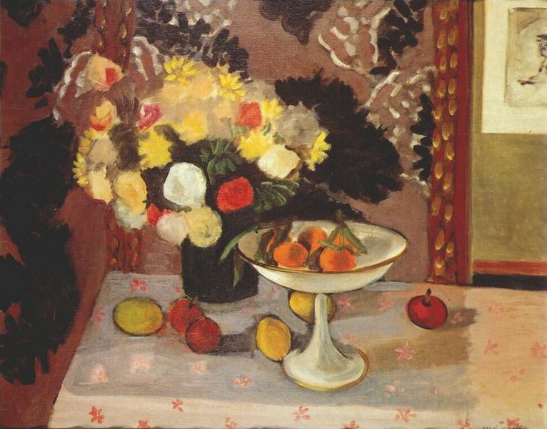 A warm interior still life by Henri Matisse with a black vase of cream, yellow, and red flowers beside a white pedestal bowl of oranges on a pale patterned cloth, set against a mauve-brown wall with dark leafy motifs and a narrow framed picture at right.