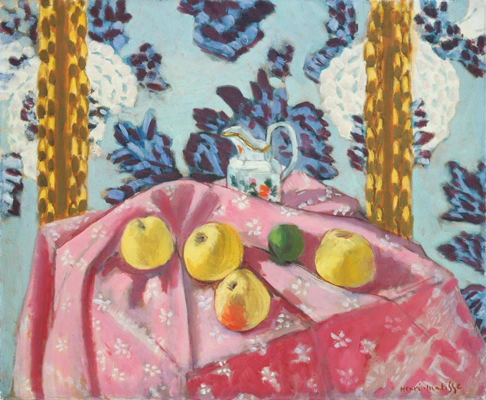 On a pink flowered tablecloth pulled into a central ridge, several yellow apples and a green citrus sit beside a small gold-rimmed pitcher; behind them a pale blue wall with dark indigo leaves and two vertical gold bands frames the bright, decorative still life.