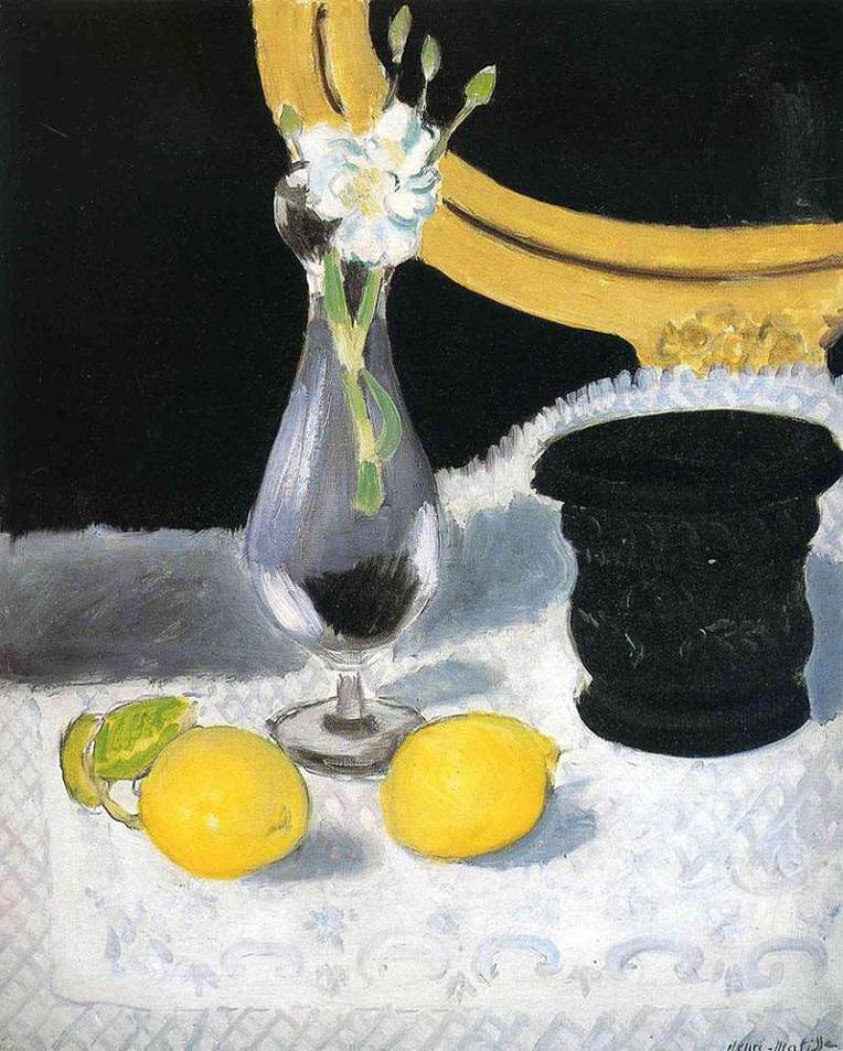 Two bright lemons rest on a white damask cloth beside a glass vase of white flowers and a matte black vessel, with a curved gold mirror against a black background anchoring the scene.