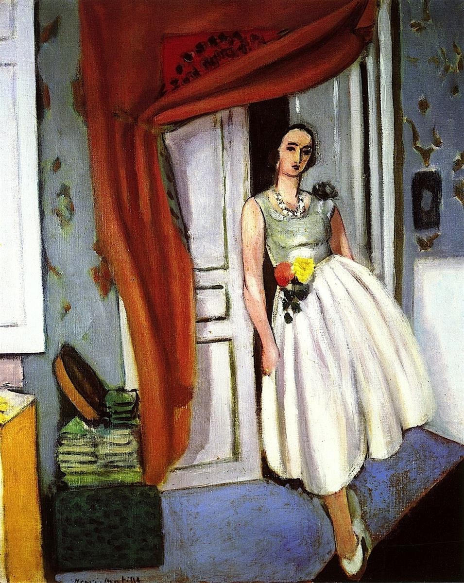 A woman in a pale dress with a floral waist corsage leans in a doorway beneath a red curtain; blue floor, patterned gray-blue walls, stacked books and a hat box at left, and soft door panels create a shallow interior stage, painted by Henri Matisse in 1926.