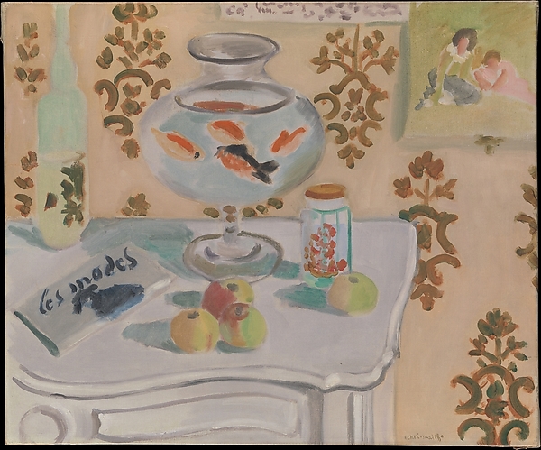Interior still life with a large glass bowl of orange and dark goldfish on a white scalloped table; nearby are apples, a green fruit, a jar with red cherries, a pale green bottle, and an angled magazine; beige wallpaper with olive arabesques and a small framed vignette hang behind, all painted in soft Nice-period tones.