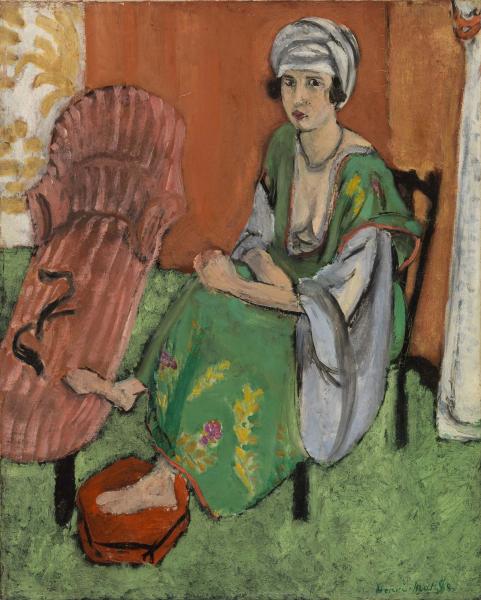 A woman in a vivid green dress and white turban sits in a shallow room with a pink chaise, a red foot basin at her bare foot, a warm terracotta wall, and a white column; loose dark contours and broad color fields characterize the painting.